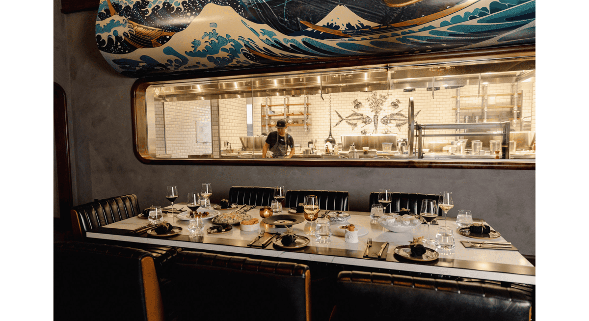 Open kitchen fine dining restaurant scene with chef preparing dishes behind glass and a table set for a luxury dining experience.
