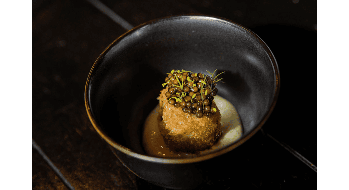 Crispy gourmet appetizer topped with caviar and microgreens, served in a black bowl with delicate sauce, showcasing refined fine dining presentation.
