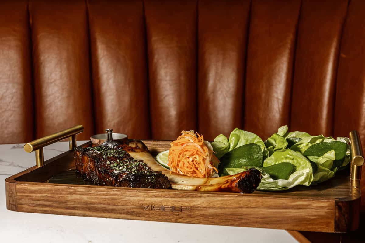 Glazed beef rib served on a wooden platter with lettuce, lime, and slaw, showcasing restaurant steak presentation and flavor-enhancing accompaniments.