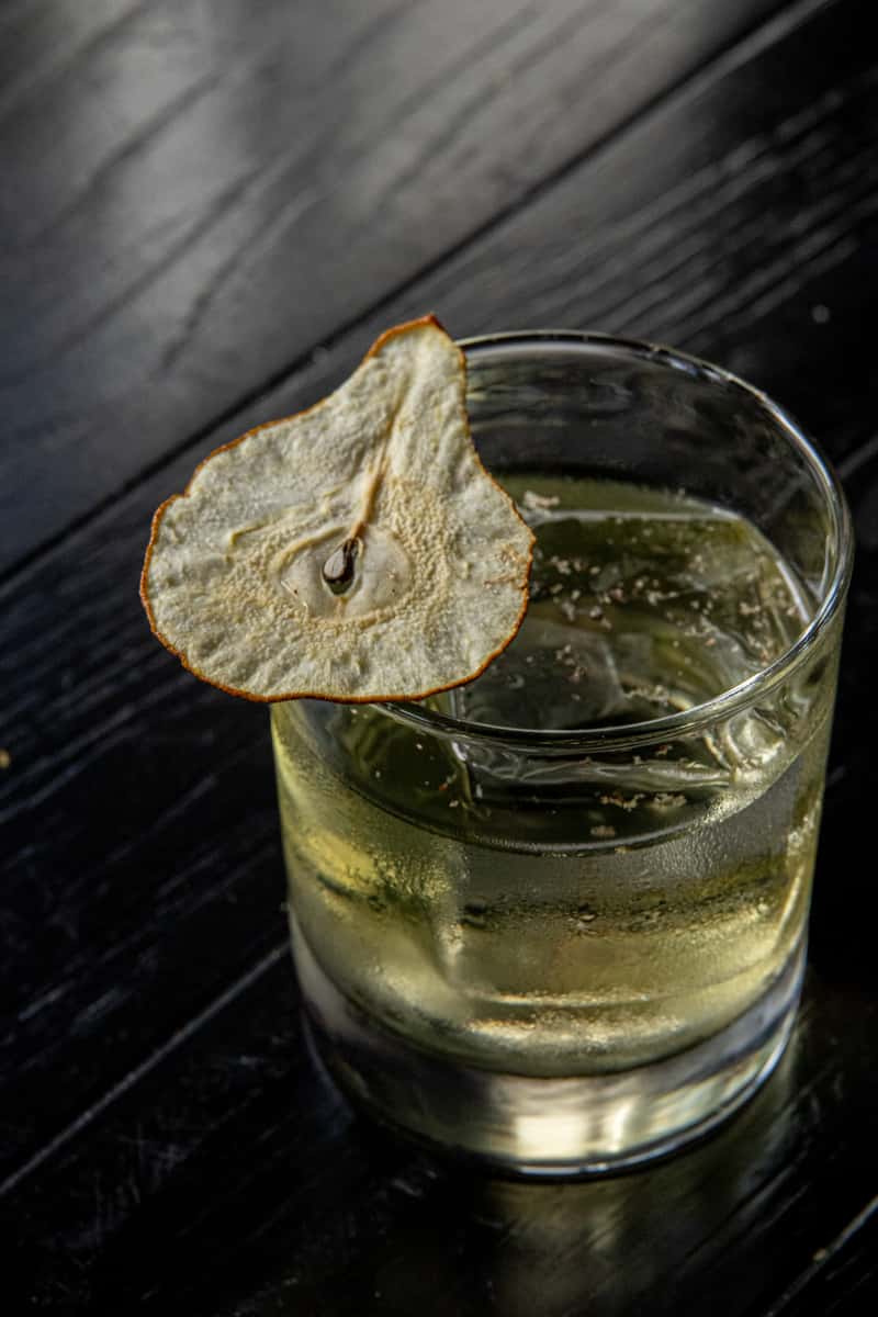 Craft cocktail with ice and dried pear garnish served on a dark bar surface