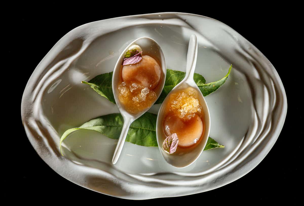 Peach dessert served on porcelain spoons with granita and fresh herb leaves.