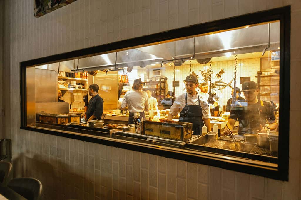 Open kitchen at a fine dining restaurant showing chefs preparing dishes behind a glass window, highlighting the interactive experience of a chef's table versus a tasting menu.