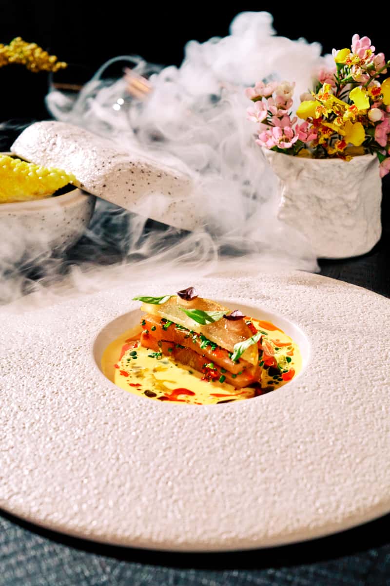 Sake and citrus-cured Japanese yellowtail plated with herbs and colorful sauce in a fine dining presentation.