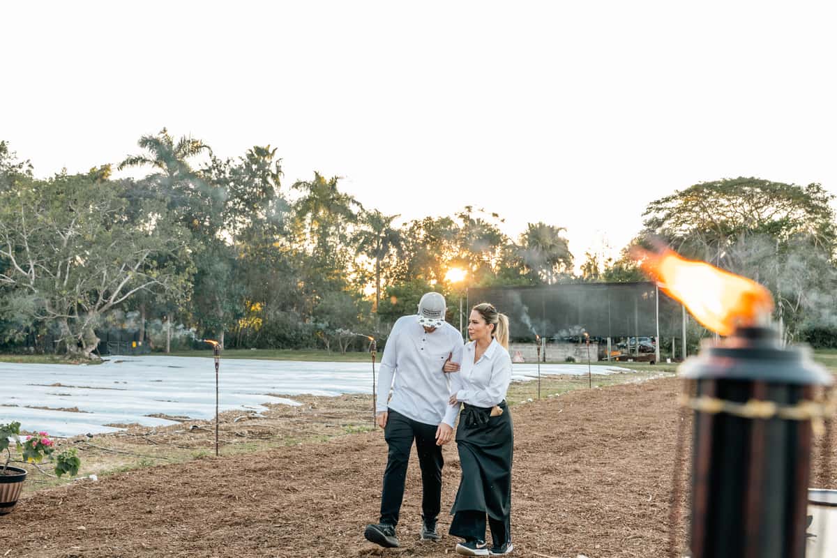 Two restaurant staff members walking through a farm field lined with torches at sunset.