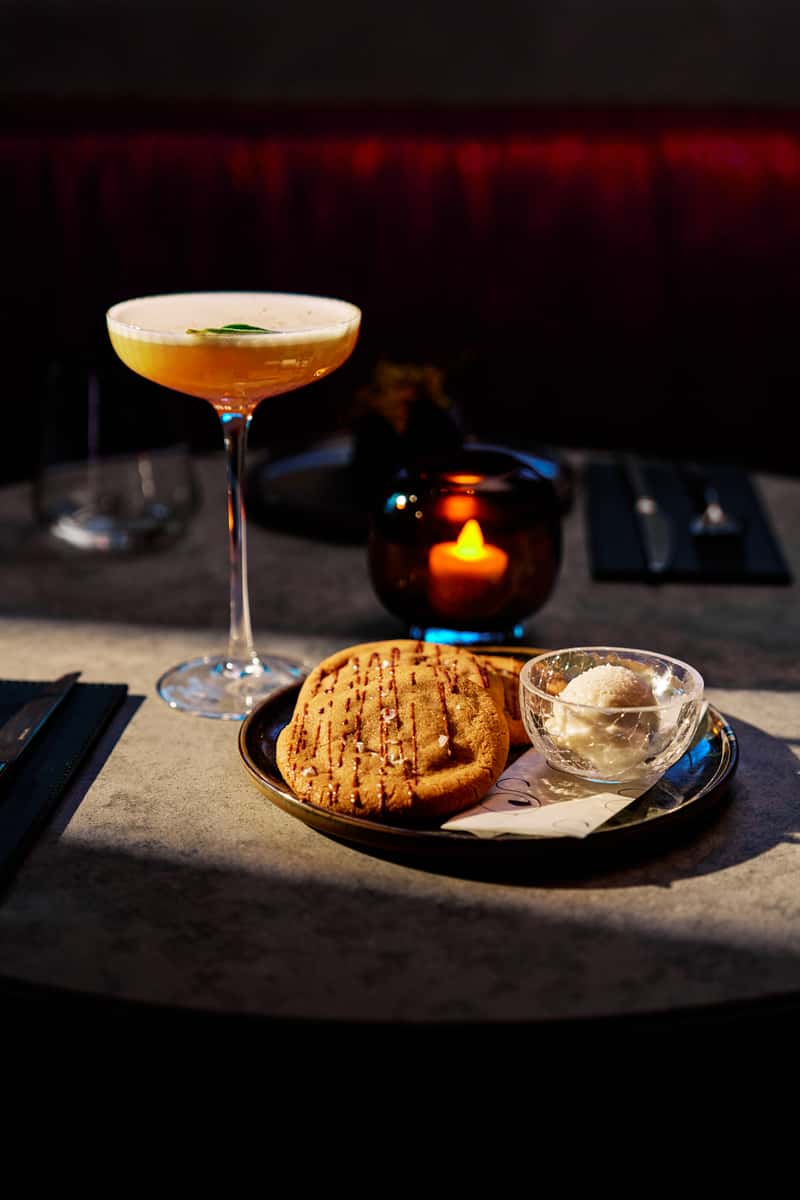 Elegant cocktail served with a warm cookie and scoop of ice cream beside candlelight.
