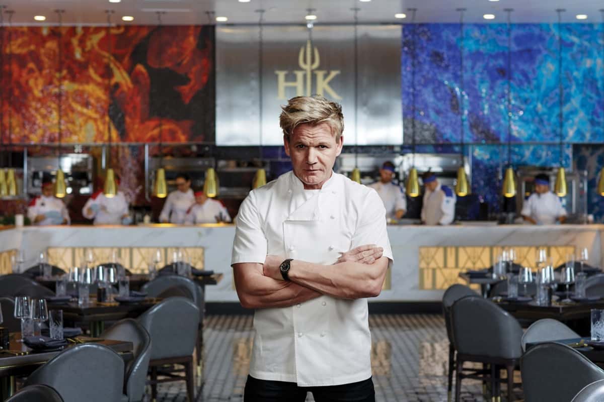 Chef Gordon Ramsay standing with arms crossed in a white chef's jacket inside Hell's Kitchen restaurant, featuring an open kitchen and elegant dining area.