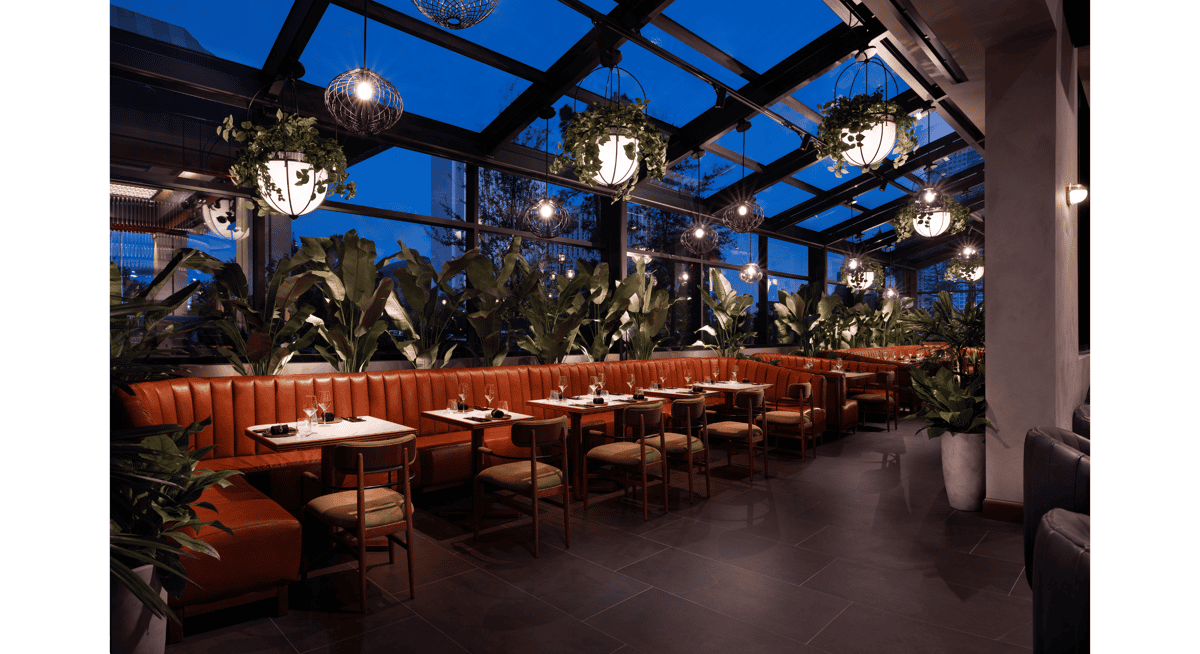 Modern upscale restaurant interior with glass ceiling, leather booth seating, hanging lights, and lush indoor plants