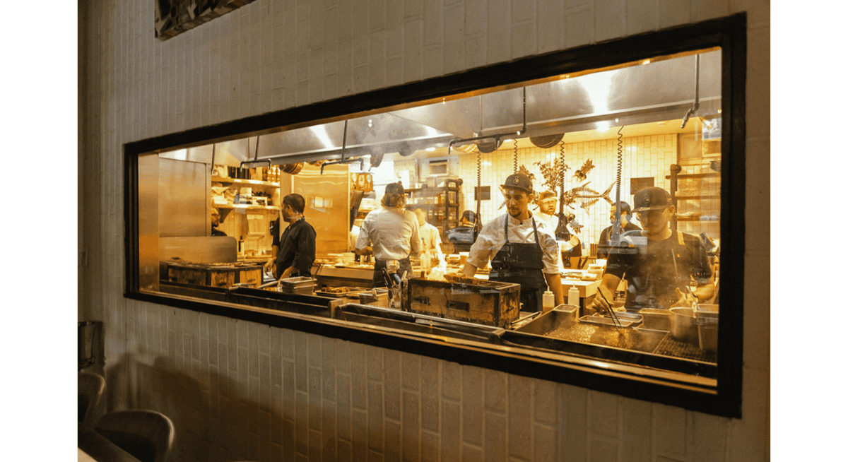 View through a restaurant window into an open kitchen where chefs prepare dishes in a busy fine dining restaurant