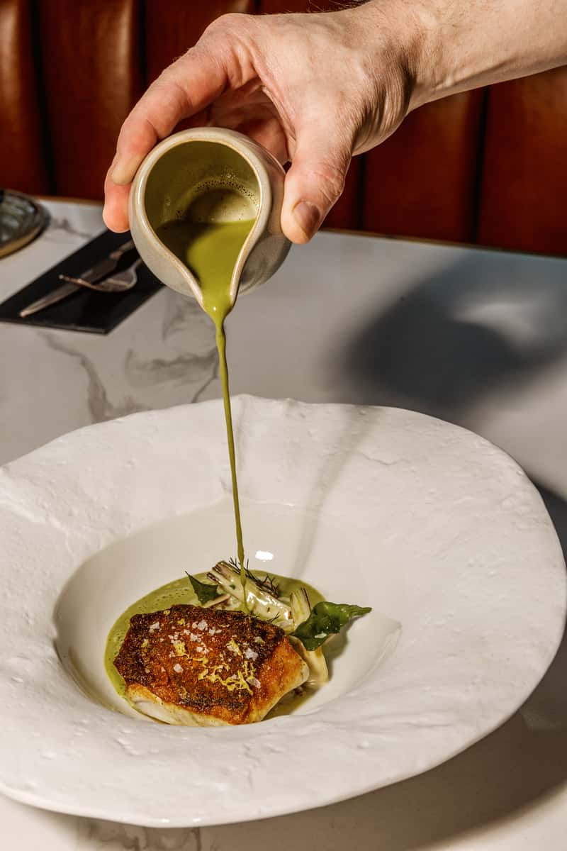Chef pouring green sauce over a fine dining fish dish at restaurant