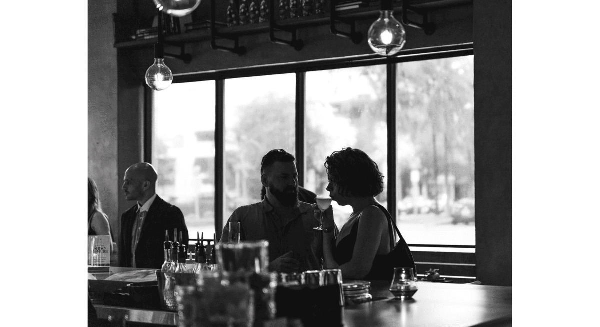 Guests enjoying drinks at a stylish restaurant bar with large windows and hanging pendant lights