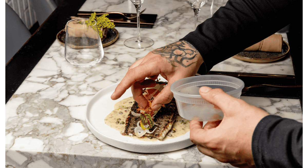 Chef plating a gourmet fish dish with vegetables and sauce on a marble countertop in a fine dining kitchen.
