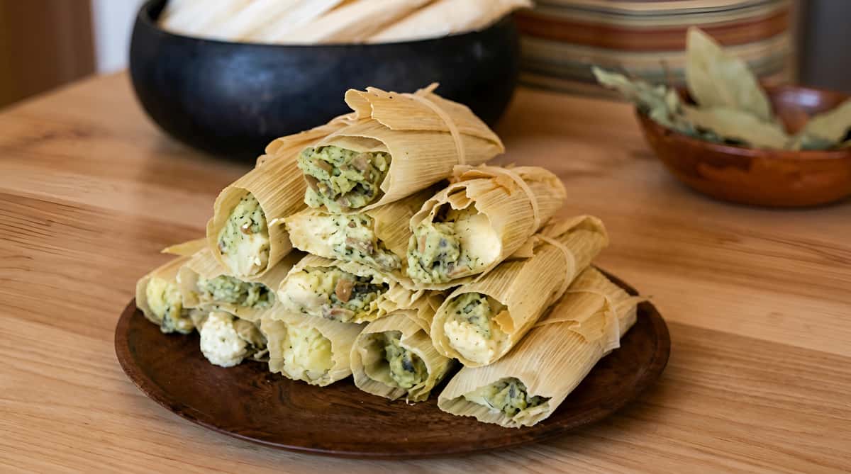 A pile of green chile and cheese tamales wrapped in corn husks on a brown plate.