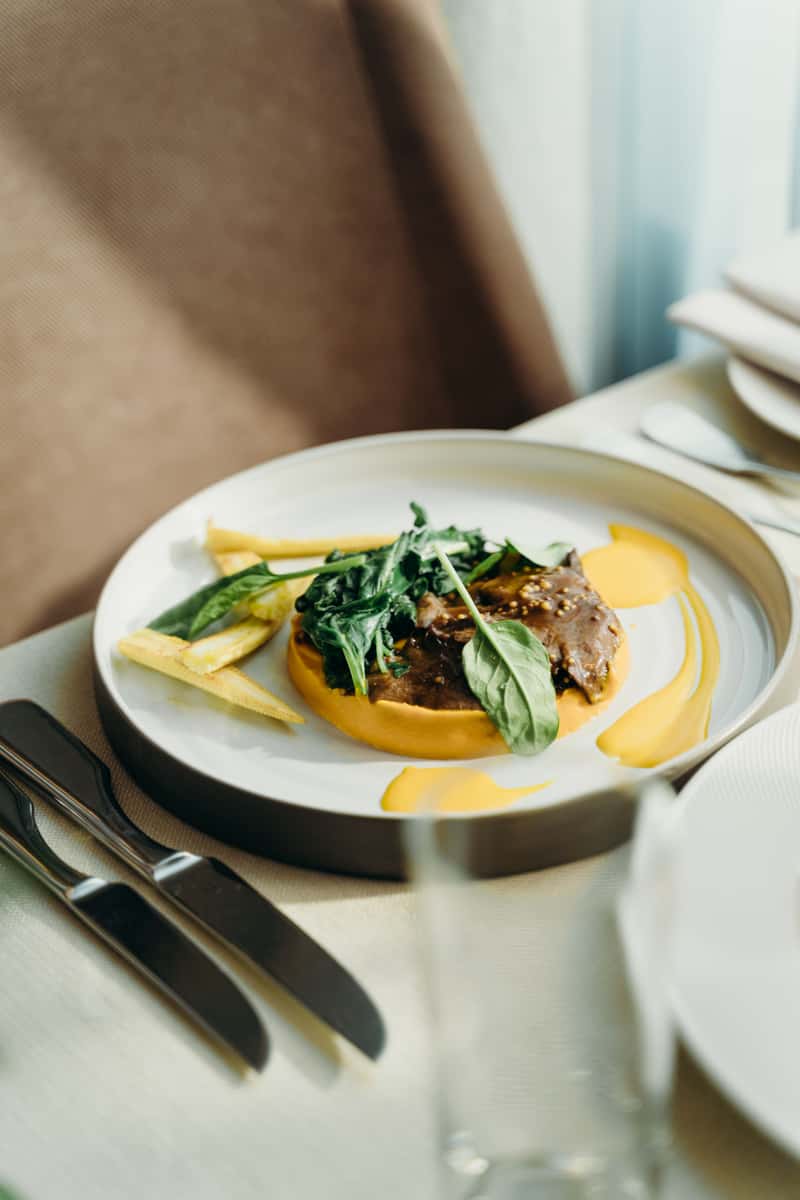 Elegantly plated gourmet dish featuring fresh green vegetables, herbs, and a golden sauce in a white ceramic bowl, served on a restaurant table.