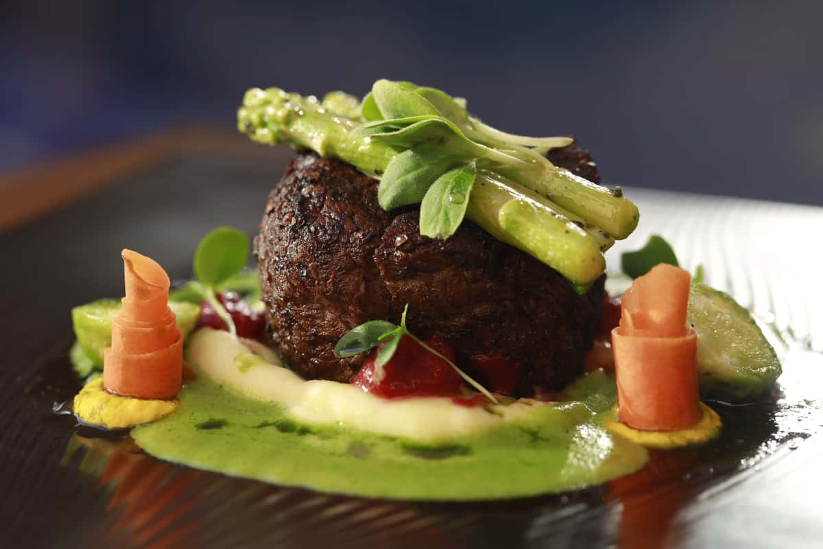 Gourmet seared beef entrée topped with fresh green vegetables, served with a vibrant green puree, roasted vegetables, and colorful garnishes on a dark plate.