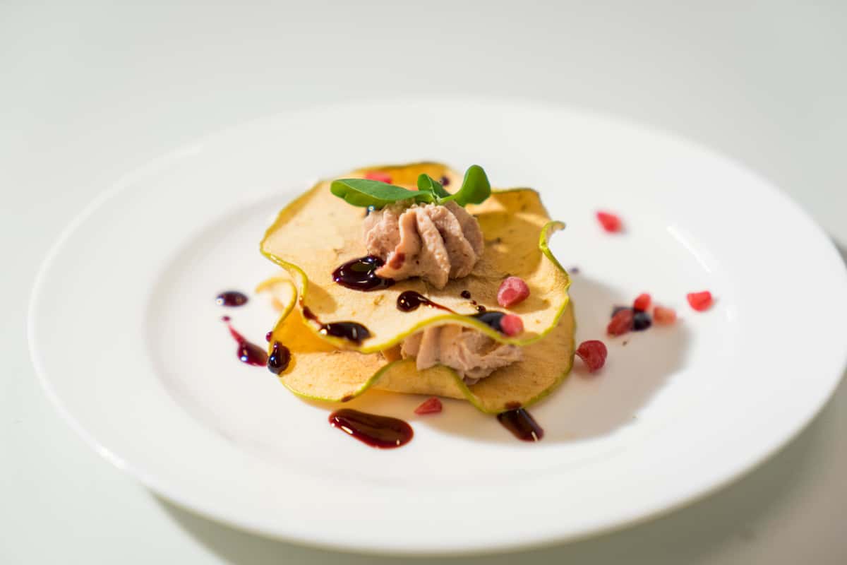 Elegantly presented gourmet dish featuring tender slices of meat layered over a golden base, garnished with fresh herbs, colorful vegetables, and decorative sauce accents on a white plate.