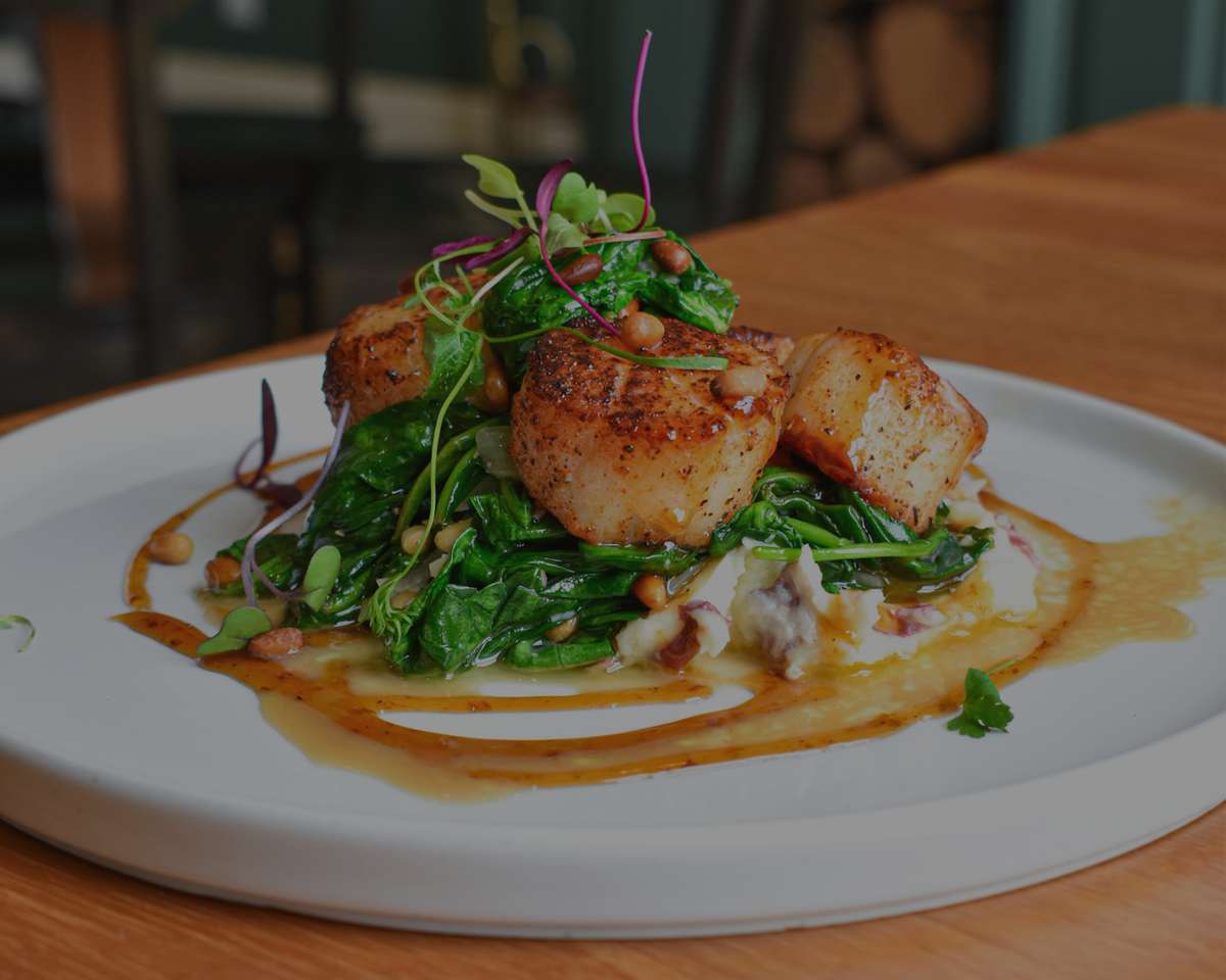 Seared scallops plated atop sautéed greens and creamy mashed potatoes, finished with toasted pine nuts, microgreens, and a golden Creole-style butter sauce. Captured inside Lou Lou on Market, this dish showcases the restaurant's bold, upscale fusion of Cajun and Mediterranean flavors.