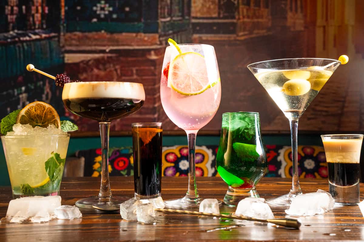 A colorful array of cocktails displayed on a wooden table, featuring a mint mojito, pink lemonade, martini with olives, and layered shots, surrounded by ice cubes.