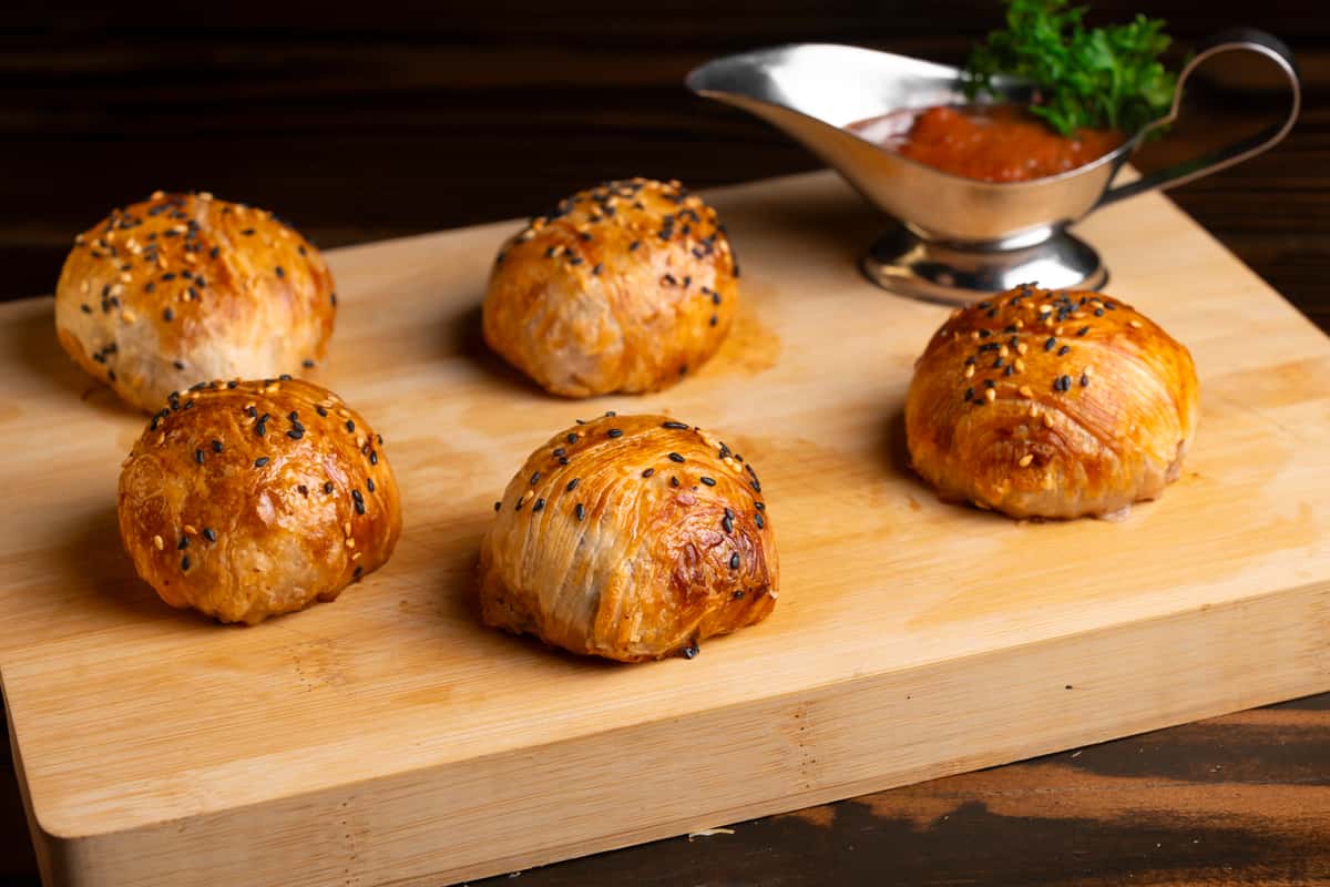 A wooden platter displays five golden-brown pastries topped with black sesame seeds, accompanied by a small silver gravy boat filled with sauce, perfect for catering events.