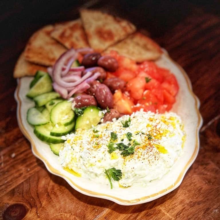Mediterranean appetizer platter with tzatziki, Kalamata olives, cucumbers, tomatoes, red onions, and warm pita bread slices.