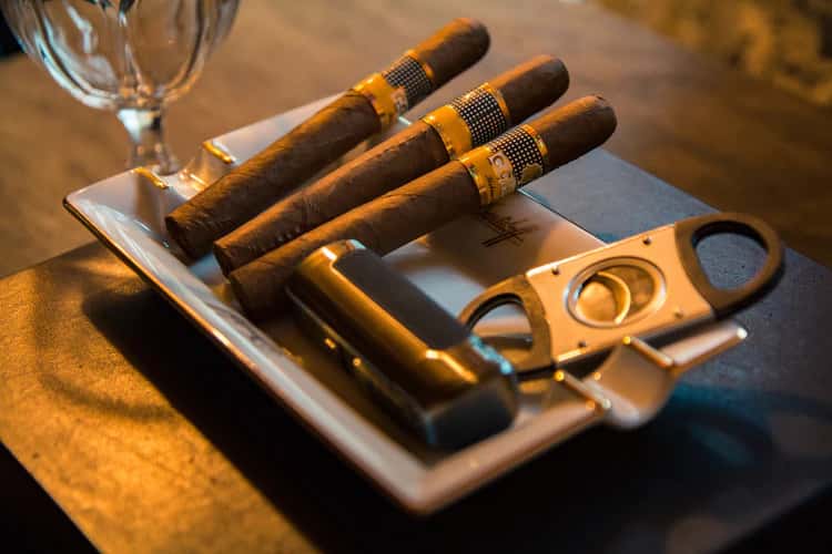 Three Cohiba cigars arranged on a rectangular tray with a cigar cutter and lighter, set on a dark tabletop beside a glass.