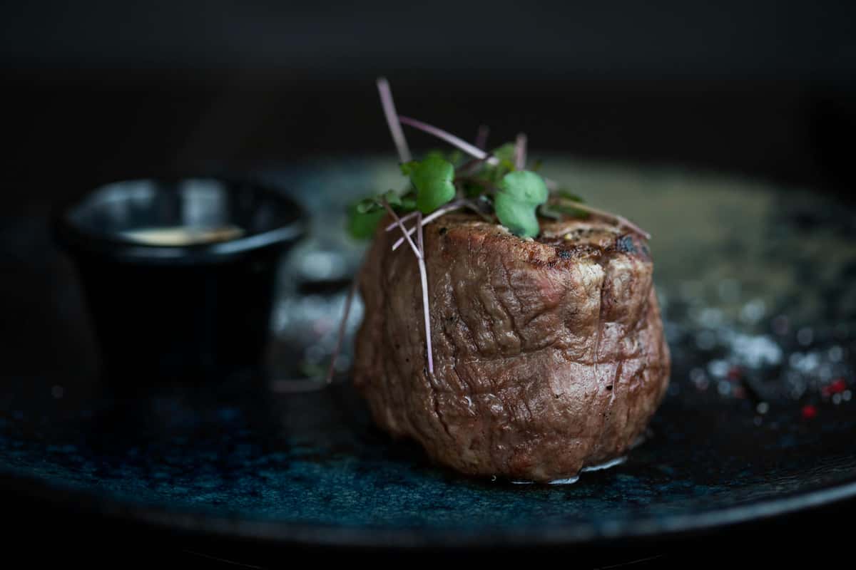 Grilled filet mignon topped with microgreens, served on a dark plate with a small black cup of creamy dipping sauce.