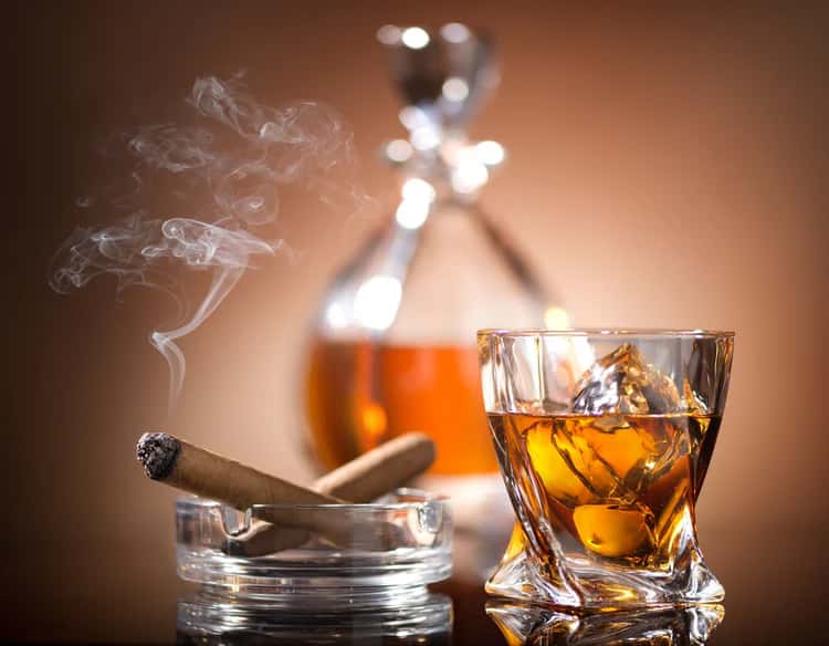 Glass of whiskey with ice next to a lit cigar in an ashtray and a decanter in the background on a warm-toned surface.