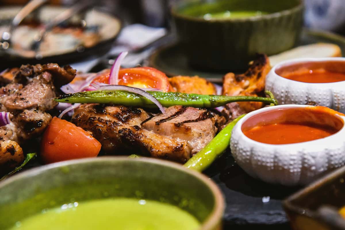 This image shows a close-up of grilled meat served with green peppers, tomato slices, and red onion, accompanied by dipping sauces—one red and one green—in small bowls. The dish appears to be a flavorful and colorful grilled platter, likely inspired by Mediterranean or Middle Eastern cuisine.