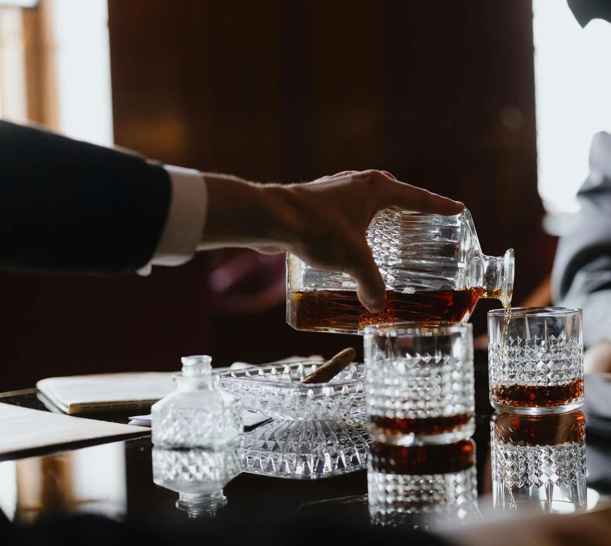 Hand pouring dark liquor from a crystal decanter into cut-glass tumblers on a reflective table.