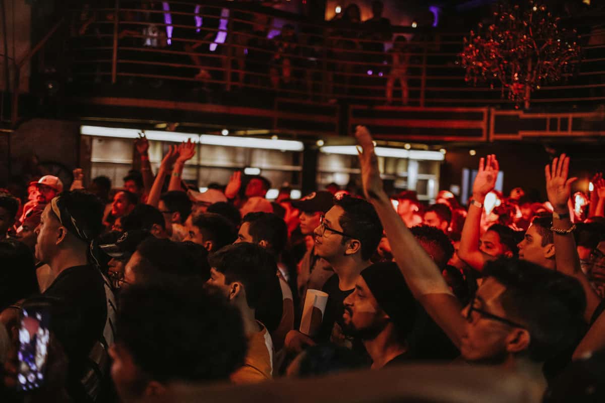 A large, diverse crowd of people dancing and raising their hands at a nightclub, lit with red and purple lights.