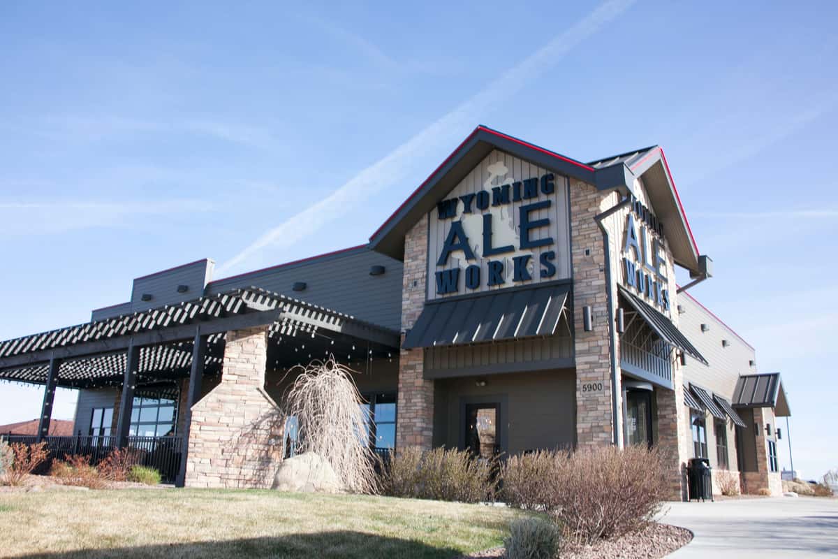 Exterior view of Wyoming Ale Works, a modern rustic building with stone accents and a large outdoor patio area under a pergola, set against a clear blue sky.