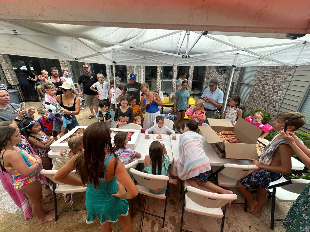 Large group of children and adults gathered under a canopy for a donut party, with open boxes of pastries on the tables