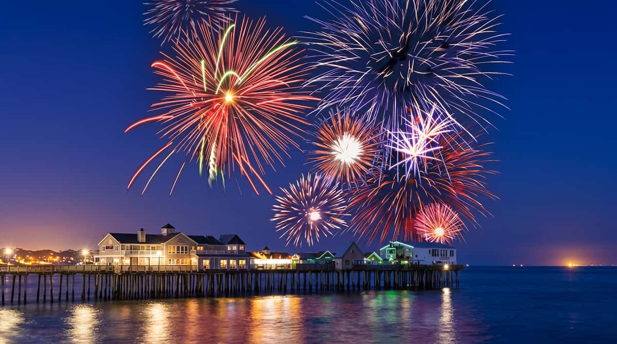 Colorful fireworks burst over a nighttime ocean pier with illuminated buildings.