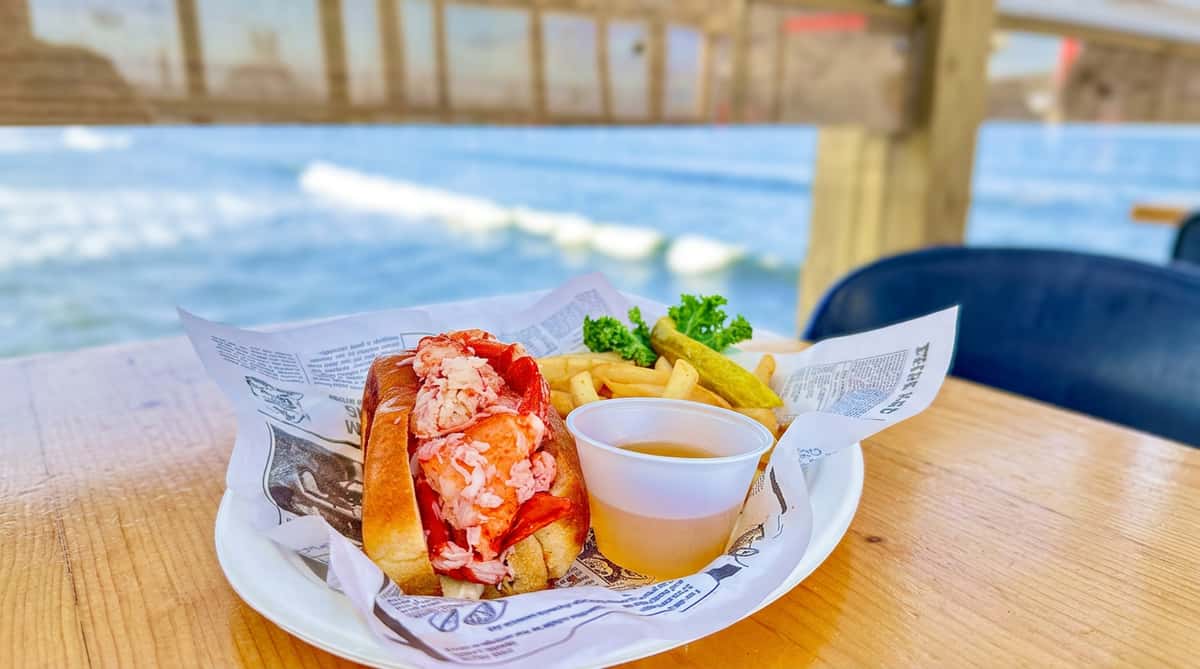 Lobster roll with fries, a pickle, and a cup of broth on a plate, overlooking the ocean.