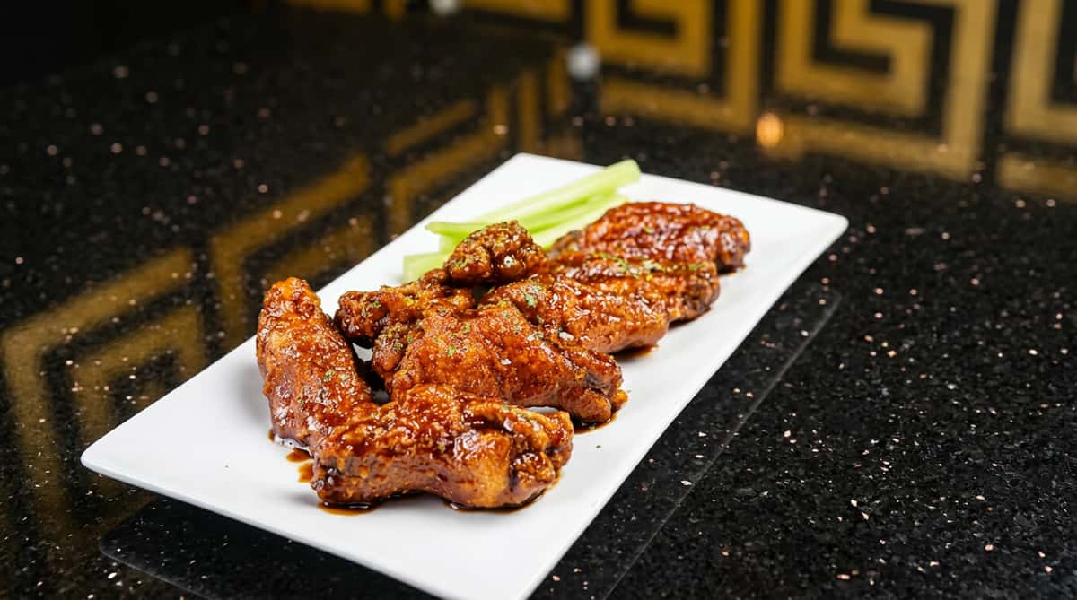 Glazed chicken wings served on a white rectangular platter with fresh celery sticks, featuring a crispy, caramelized coating in a modern restaurant setting.