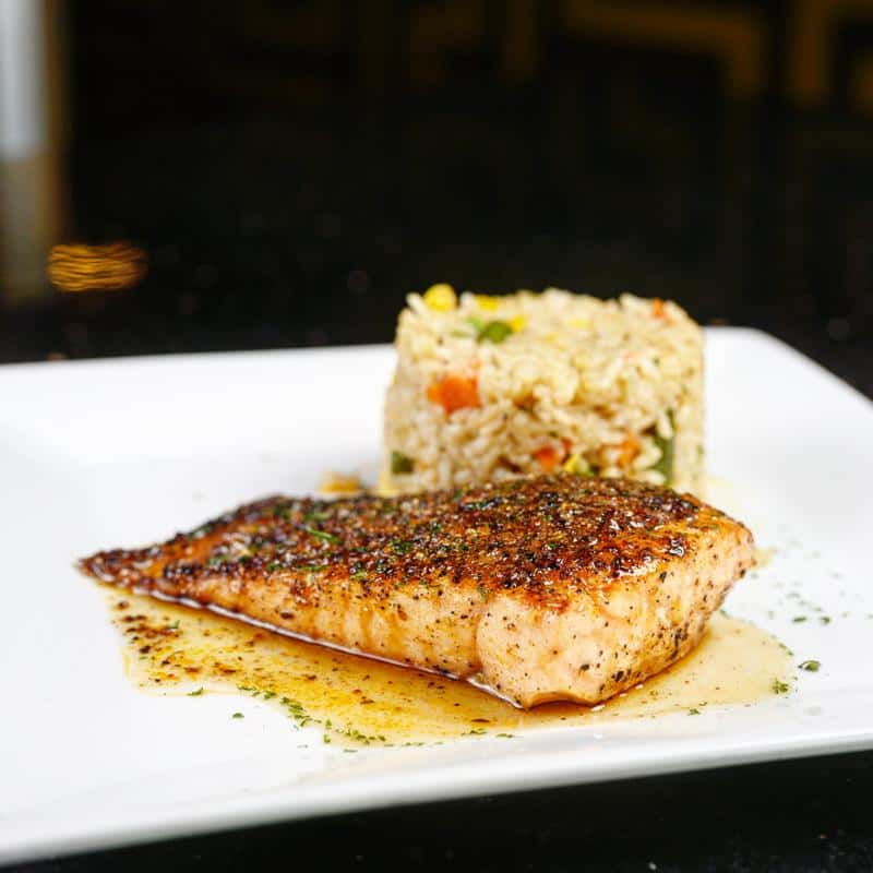 Blackened salmon fillet with seasoned rice pilaf on a white plate, drizzled with buttery herb sauce.