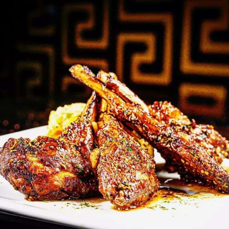 Glazed lamb chops stacked on a white plate, served with a side of seasoned rice and garnished with herbs.