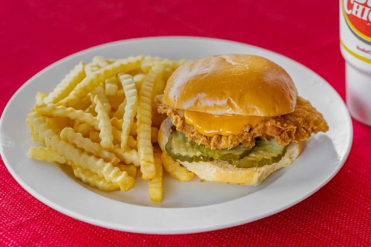 Crispy chicken sandwich with cheddar cheese and pickles on a glossy toasted bun, served alongside crinkle-cut fries on a white plate over a red textured tablecloth.