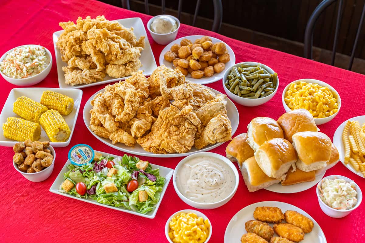 Hearty Southern comfort food feast featuring crispy fried chicken, chicken tenders, fluffy dinner rolls, hush puppies, crinkle-cut fries, mac and cheese, green beans, coleslaw, corn on the cob, fried okra, a fresh garden salad with buttermilk ranch, and creamy white gravy, all beautifully arranged on a vibrant red tablecloth.