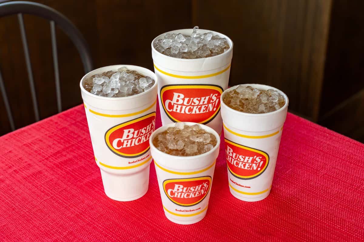 Assortment of iced soft drinks in various sizes served in Bush's Chicken branded cups, displayed on a red textured tablecloth inside a casual dining setting.