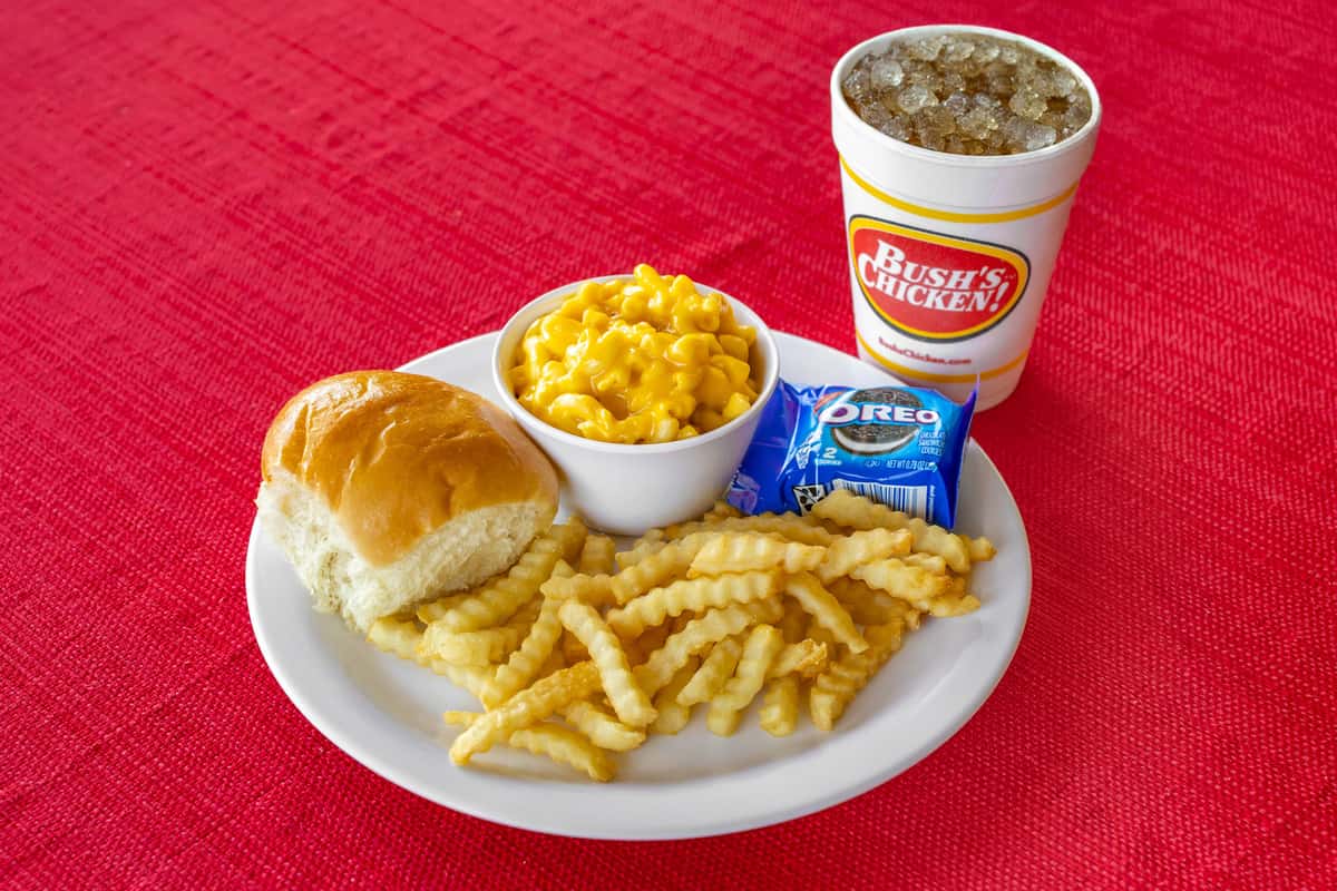 Kid's meal from Bush's Chicken featuring crinkle-cut fries, creamy mac and cheese, a soft dinner roll, a pack of Oreo cookies, and a large iced soft drink, all served on a white plate over a bright red tablecloth.