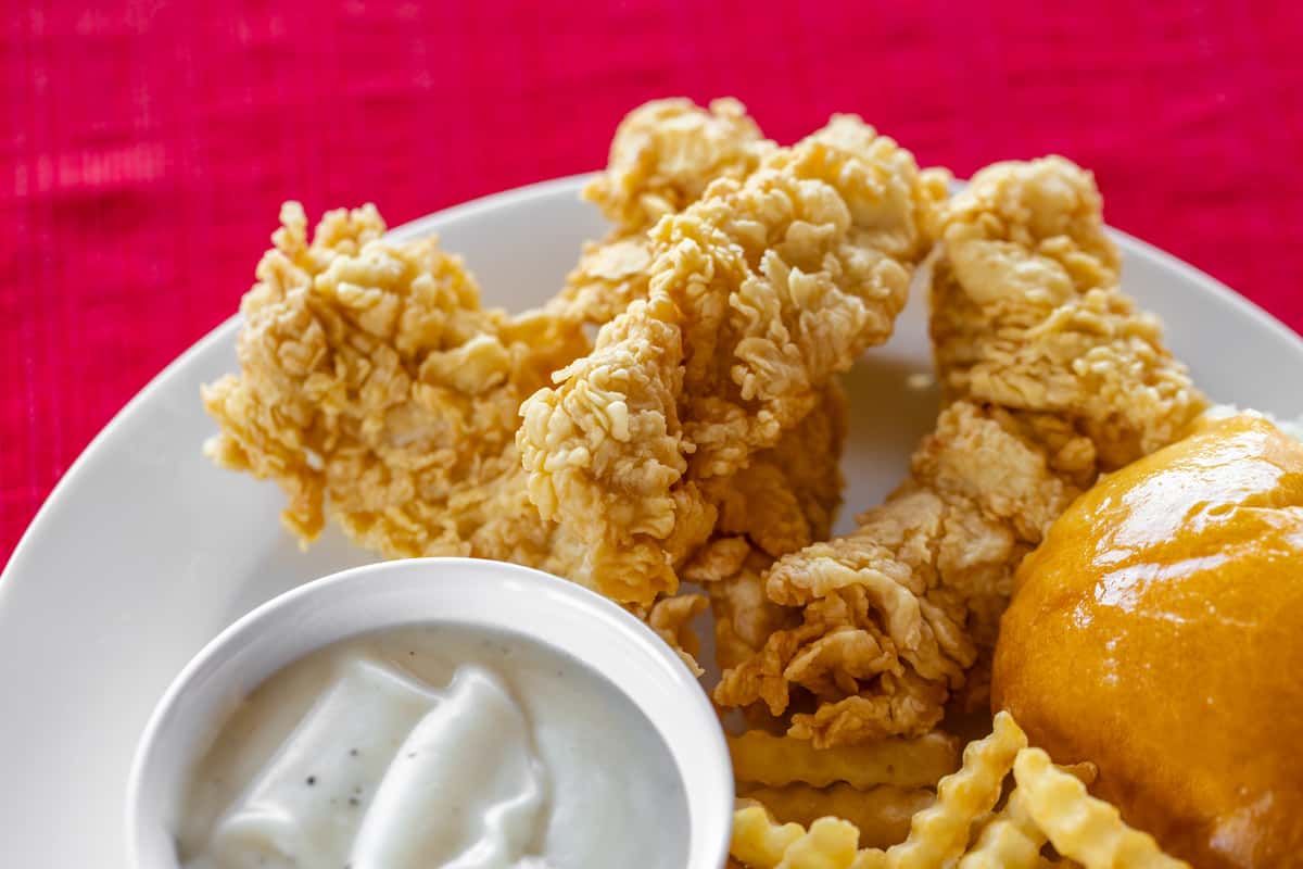Close-up of Bush's Chicken crispy chicken tenders served with crinkle-cut fries, creamy white gravy, and a golden dinner roll on a white plate with a vibrant red tablecloth backdrop.