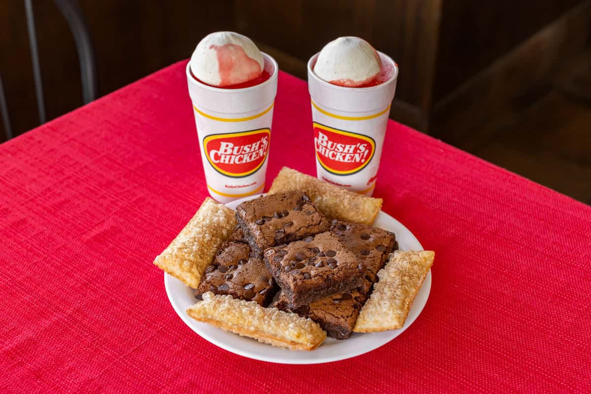 Plate of chocolate chip brownies and crispy fried dessert pies served with two Bush's Chicken strawberry floats topped with vanilla ice cream, displayed on a vibrant red tablecloth.