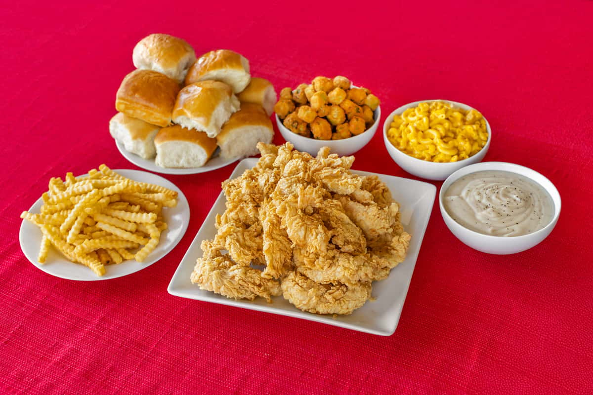Southern-style combo meal featuring a generous plate of crispy chicken tenders, crinkle-cut fries, fluffy dinner rolls, fried okra, creamy mac and cheese, and a bowl of peppered white gravy, all set on a vibrant red tablecloth for a classic comfort food presentation.