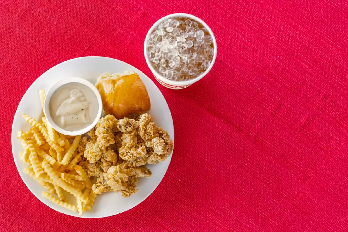 Crispy chicken gizzards with crinkle-cut fries, a fluffy dinner roll, creamy white gravy, and a large iced drink served on a white plate atop a bold red tablecloth at Bush's Chicken.