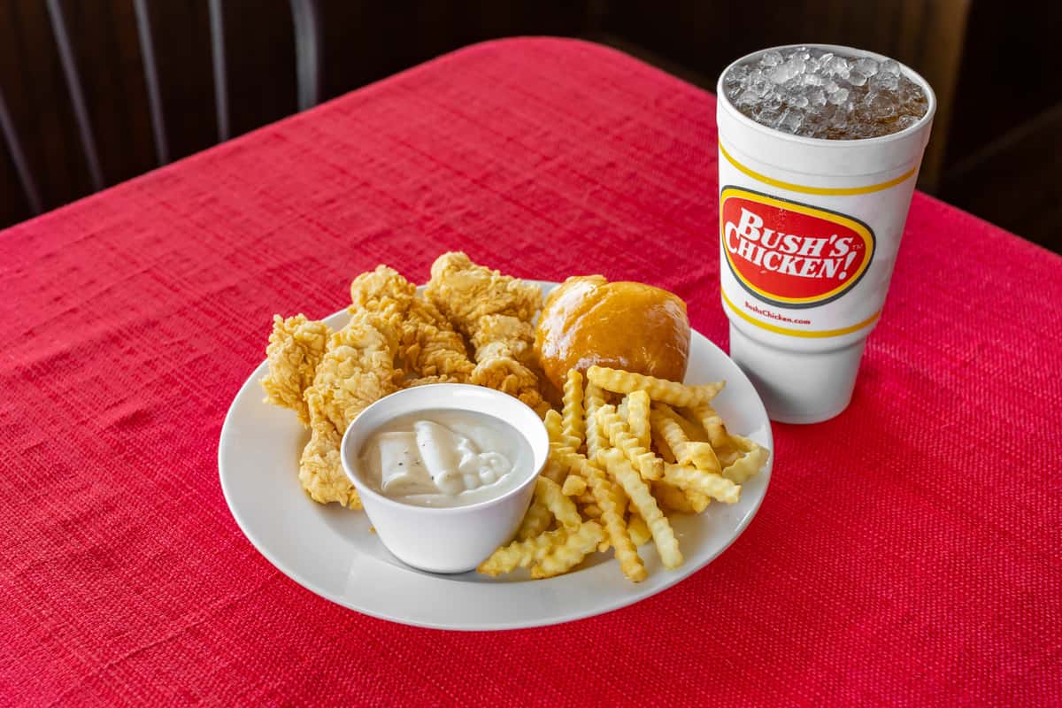 Classic Southern meal from Bush's Chicken featuring crispy chicken tenders, crinkle-cut fries, a fluffy dinner roll, and a cup of creamy white gravy, served on a white plate with an iced soft drink on a red tablecloth.