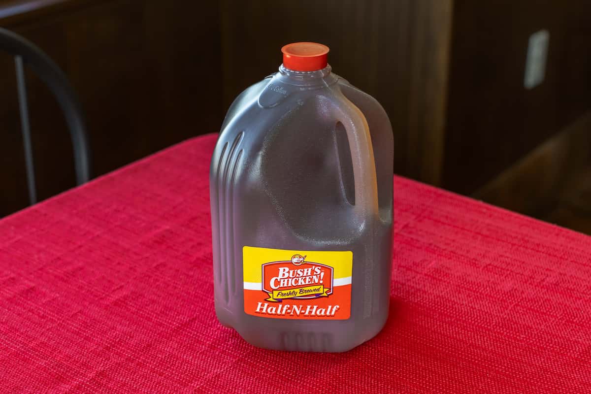 One-gallon jug of Bush's Chicken Half-N-Half tea, featuring a mix of sweet and unsweet iced tea, displayed on a red tablecloth in a casual dine-in setting.