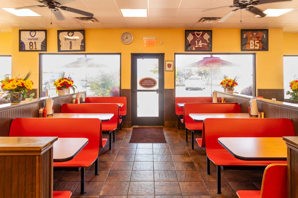 Interior of a Bush's Chicken restaurant featuring bright red booth seating, sports memorabilia on yellow walls, autumn-themed floral arrangements, and a welcoming atmosphere with natural light streaming through large front windows.