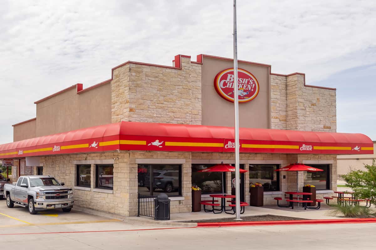 Exterior of a Bush's Chicken restaurant featuring tan brick walls, a bold red awning with yellow accents, outdoor seating with red umbrellas, and a pickup truck parked in front under a partly cloudy sky.