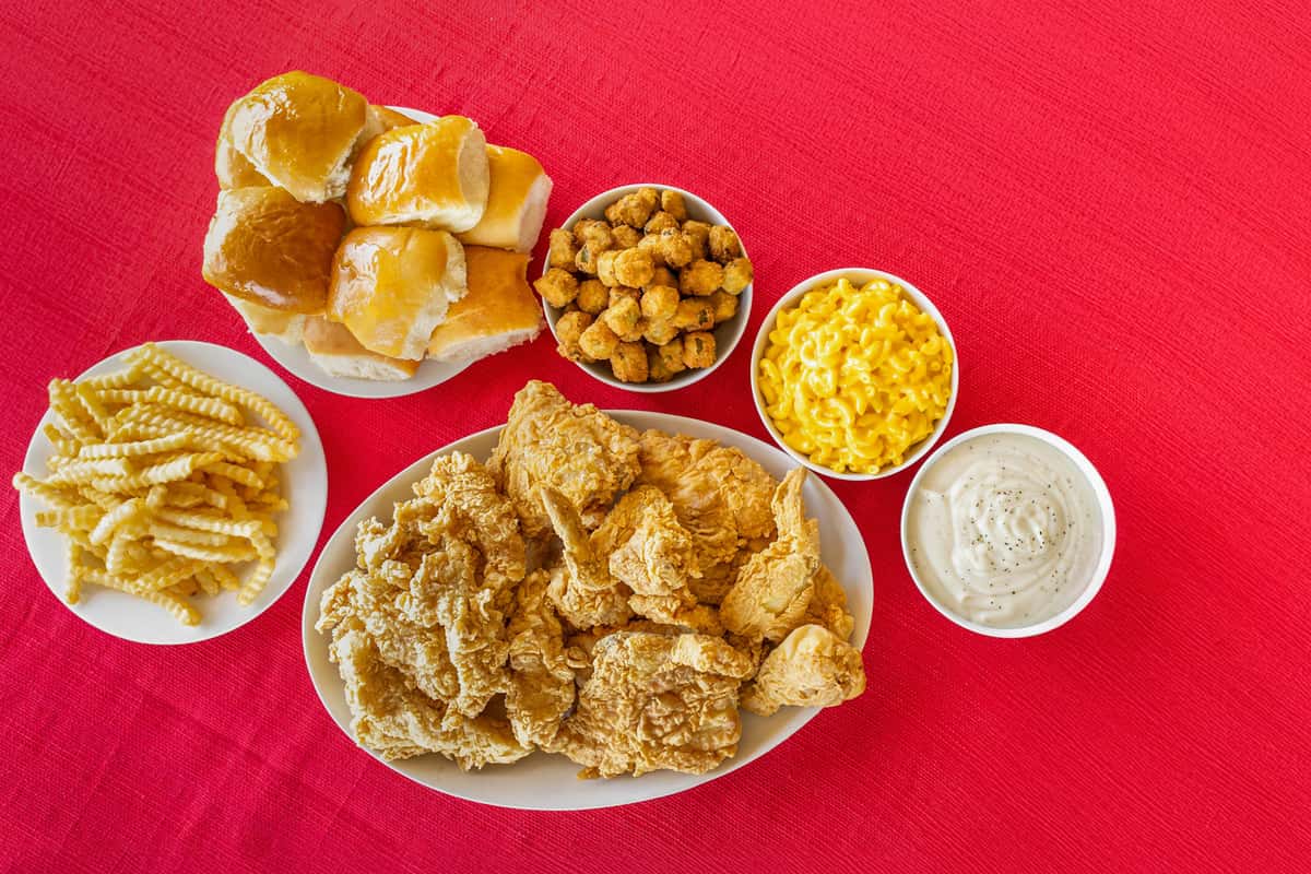 Southern-style comfort meal featuring a large platter of crispy fried chicken, crinkle-cut fries, fluffy golden dinner rolls, fried okra, creamy mac and cheese, and a bowl of white peppered gravy, all arranged on a red textured tablecloth.