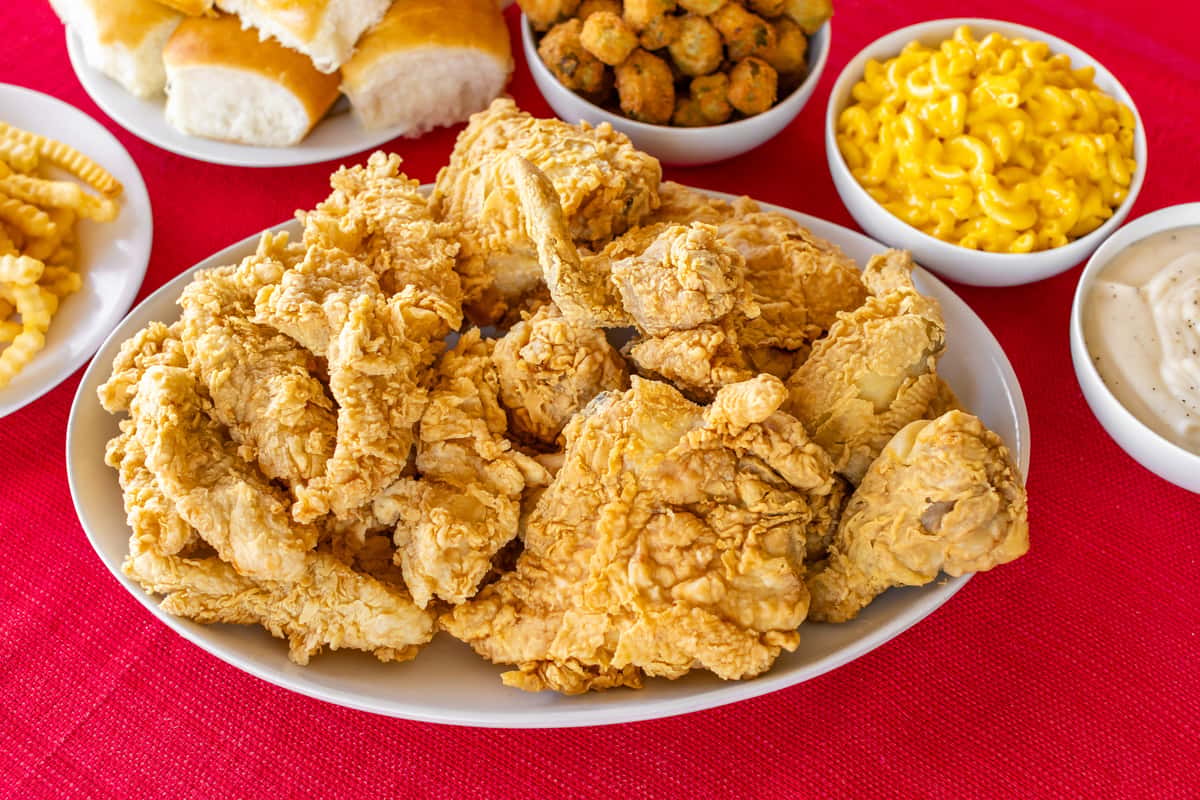 Southern-style fried chicken platter with crispy golden-brown pieces, surrounded by classic comfort food sides including creamy mac and cheese, fried okra, fluffy dinner rolls, crinkle-cut fries, and peppered white gravy, all served on a vibrant red tablecloth.